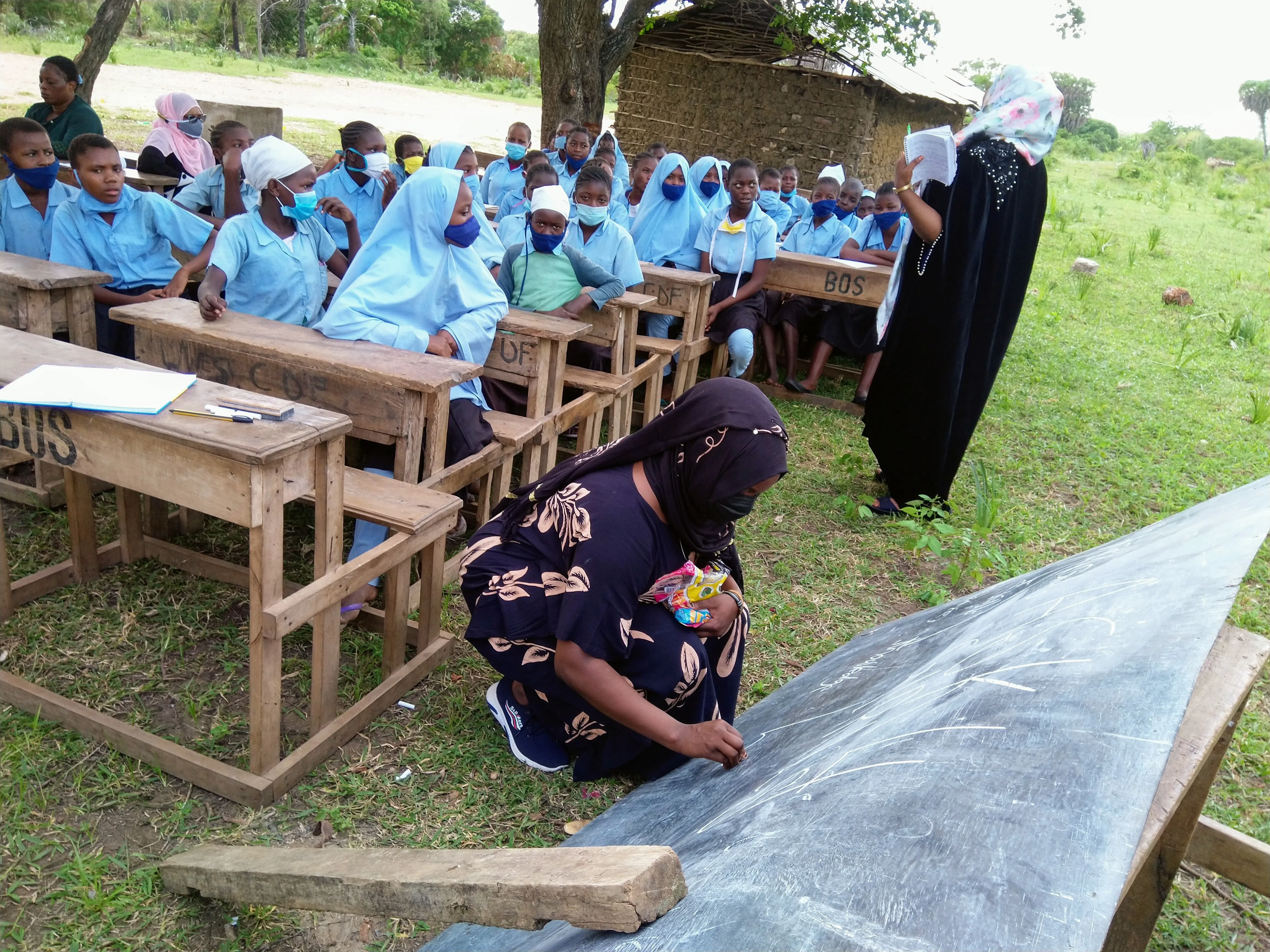 A group of kenyan girls in an outdoor classroom with their teacher. A girl is writing on a blackboard